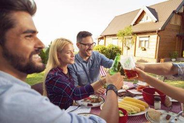 Bir grup genç arkadaş dışarıda barbekü yapıyor, masada otururken kadeh kaldırıyor, bira içiyor ve eğleniyorlar.