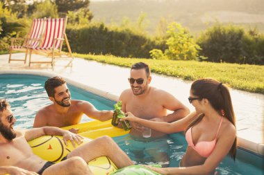 Bir grup insan havuz partisinde eğleniyor, kokteyl ve bira içiyor ve kadeh kaldırıyor.