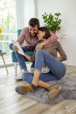 Güzel bir çift evde dinleniyor, bir adam koltukta oturuyor, tablet bilgisayar kullanıyor ve kadın yerde oturup dergi okuyor.