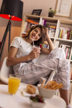 Güzel genç bayan hafta sonu sabahının tadını çıkarıyor, kahvaltı yapıyor ve taze çilekli smoothie içiyor.