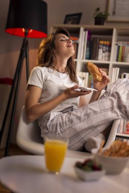 Güzel genç bir kadın pencerenin yanındaki sandalyeye oturmuş kahvaltıda taze kruvasan ve bir bardak portakal suyu içiyor.