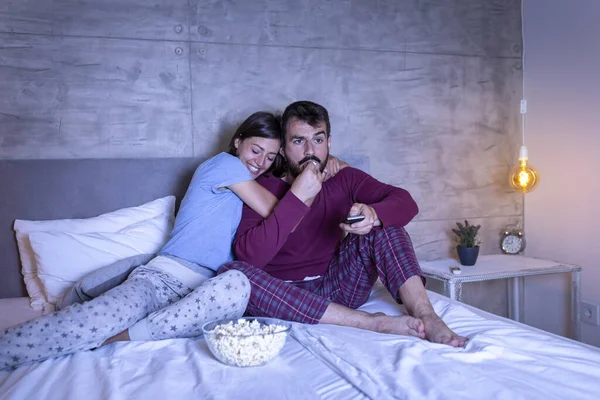 Beautiful young couple in love wearing pajamas sitting and hugging in bed, eating popcorn and watching TV at night