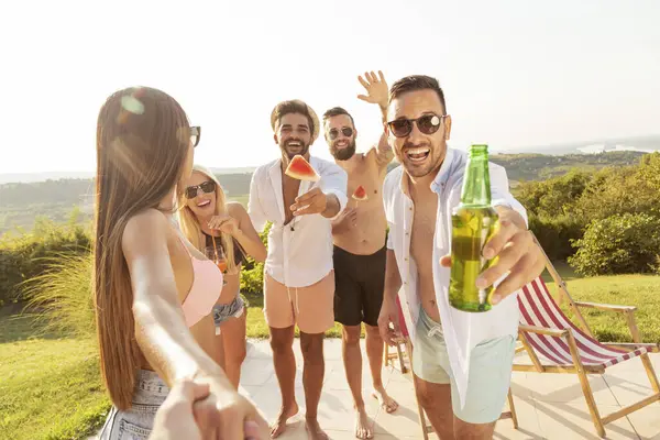 Group of young friends having fun at a poolside summertime party, drinking beer and inviting more friends to join them