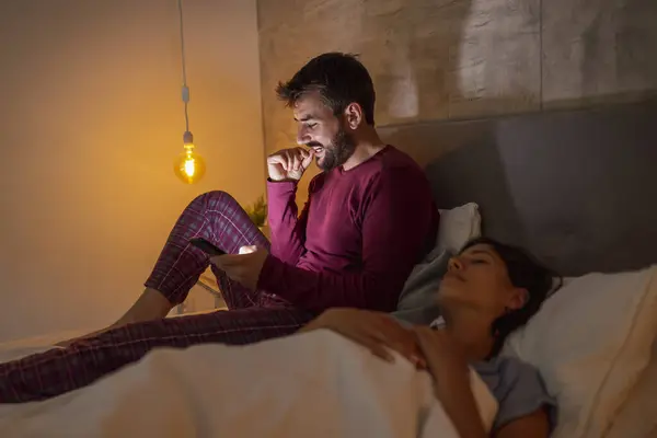 Man using smart phone while his girlfriend is asleep in bed at night; insomnia and sleeping disorders concept