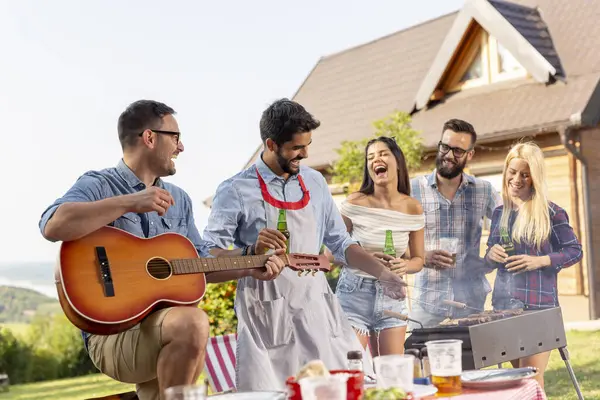 Group of friends having a barbecue party outdoors, drinking beer, grilling meat, playing the guitar and having fun