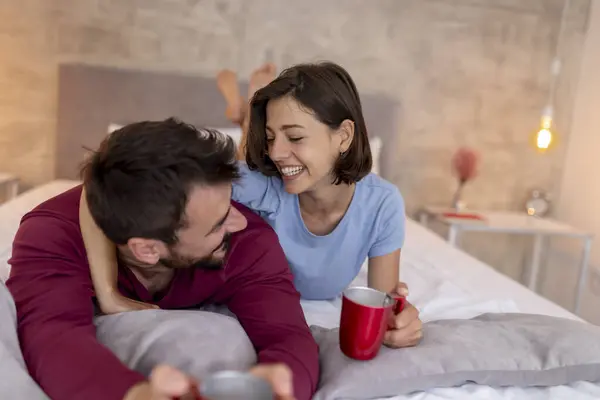 Beautiful couple in love wearing pajamas, having coffee in bed in the morning after waking up. Couple drinking coffee and having fun in bed