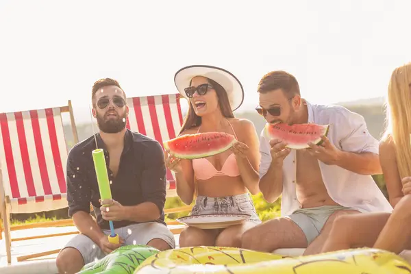 Group of friends at a poolside summer party, sitting at the edge of a swimming pool, eating cold watermelon slices and having fun