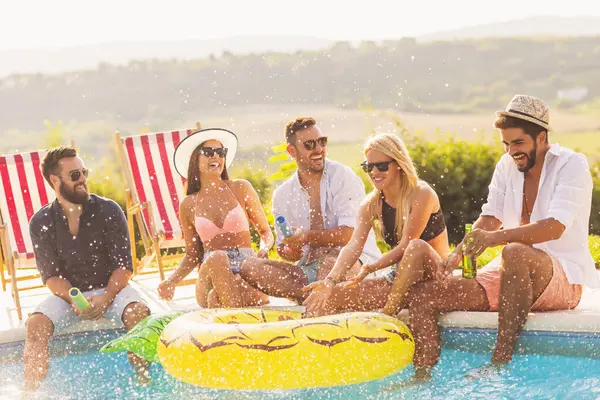 Group of friends at a poolside summer party, sitting at the edge of a swimming pool, drinking beer, splashing water and having fun