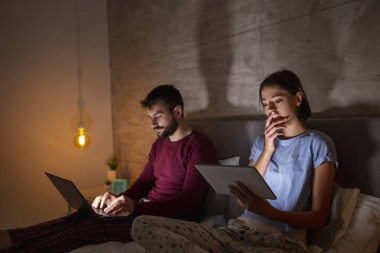 Güzel genç çift geceleri yan yana oturup laptopta ve tablet bilgisayarlarda geç saatlere kadar çalışıyorlar.