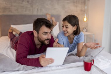 Güzel bir çift yatakta uzanıyor, kahve içiyor ve tablet bilgisayarda internette sörf yapıyor, ev dekorasyonu fikirleri arıyor.