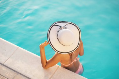 Bikini ve şapka giyen çekici genç bir kadın, yüzme havuzunda duruyor, güneşleniyor ve dinleniyor.