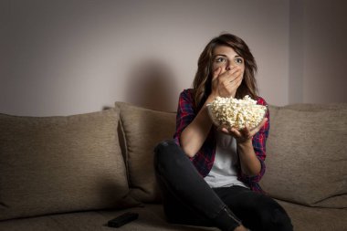 Güzel genç bir kadın oturma odasındaki kanepede oturuyor, korkmuş, korku filmi seyrediyor ve patlamış mısır yiyor.