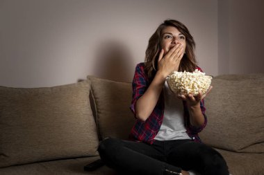 Güzel genç bir kadın, oturma odasındaki kanepede oturmuş televizyon izlerken ve patlamış mısır yerken eğleniyor.
