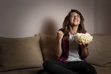 Genç ve güzel bir kadın oturma odasındaki kanepede oturuyor, TV 'de futbol maçı seyrediyor ve patlamış mısır yiyor.