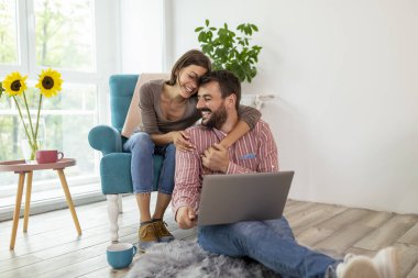 Aşık bir çift yerde oturuyor, kahve içiyor ve dizüstü bilgisayarında ev dekorasyonu fikirleri arıyor.