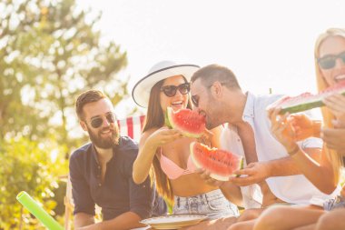 Havuz başında bir yaz partisinde bir grup arkadaş, yüzme havuzunun kenarında oturmuş, soğuk karpuz dilimleri yiyor ve eğleniyorlar.