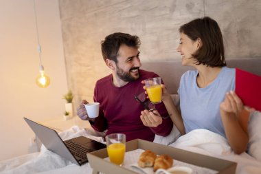 Güzel, genç ve aşık bir çift yatakta kahvaltı ederken internette kitap okuyup sörf yaparken eğleniyor.