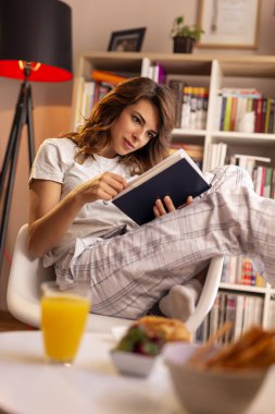 Güzel genç bir kadın pijama giyiyor, sandalyede oturuyor ve kahvaltı ederken kitap okuyor.