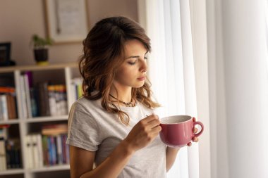 Pijama giyen, oturma odasının penceresinin yanında duran, elinde bir kase kahvaltı gevreği tutan ve hafta sonunun tadını çıkaran güzel genç bir kadın.