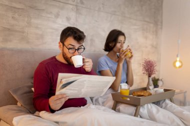 Güzel genç bir çift sabahları yatakta uzanıyor, kahvaltı yapıyor ve gazete okuyor.