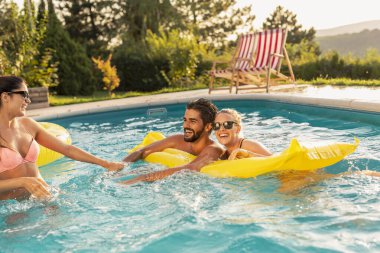 Bir grup arkadaş havuz başında bir yaz partisinde, yüzme havuzunda eğleniyorlar, yüzen yatakta güneşleniyorlar.