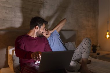 Çift geceleri yatakta yatar, dizüstü bilgisayar kullanır ve evde dinlenir, kadın yorgun ve uykulu, esniyor ve esniyor.