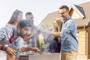 Bir grup arkadaş arka bahçede barbekü partisi veriyor, et pişiriyor, bira içiyor ve güneşli bir yaz gününde kadeh kaldırıyor.