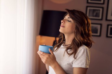 Pijama giyen güzel genç bir kadın, oturma odasının penceresinin yanında duruyor, elinde bir fincan kahve tutuyor ve anın tadını çıkarıyor.