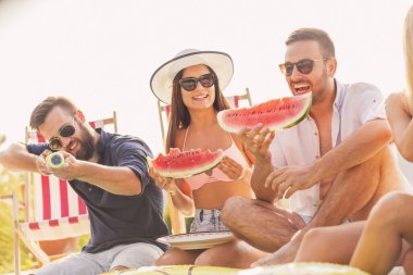 Havuz başında bir yaz partisinde bir grup arkadaş, yüzme havuzunun kenarında oturmuş, soğuk karpuz dilimleri yiyor ve eğleniyorlar.