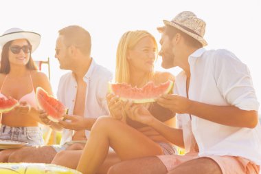 Havuz başında bir yaz partisinde bir grup arkadaş, yüzme havuzunun kenarında oturmuş, soğuk karpuz dilimleri yiyor ve eğleniyorlar.