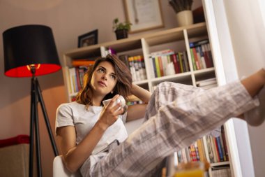 Pijama giyen, oturma odasının penceresinin yanında oturan ve sabah kahvesinin tadını çıkaran genç ve güzel bir kadın.