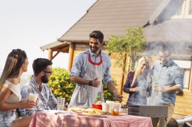 Bir grup arkadaş arka bahçede barbekü yapıyor, bira içiyor ve güneşli bir yaz gününde eğleniyorlar. Ortadaki adama odaklan.