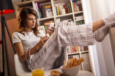 Güzel, genç bir kadın pencerenin yanında oturuyor, kahvaltı yapıyor, kahve içiyor, elinde akıllı bir telefon tutuyor ve mesaj yolluyor.