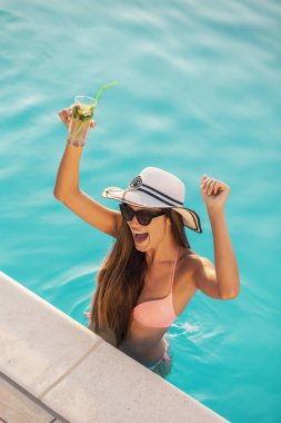 Bikini giyen, güneş gözlüğü takan ve şapka takan çekici genç bir kadın, yüzme havuzunun kenarında duruyor, kokteyller içiyor ve dans ediyor.
