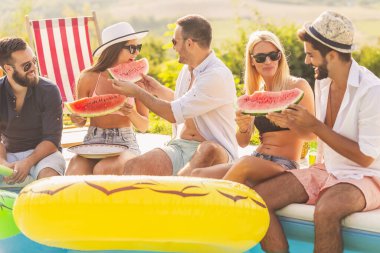 Havuz başında bir yaz partisinde bir grup arkadaş, yüzme havuzunun kenarında oturmuş, soğuk karpuz dilimleri yiyor ve eğleniyorlar.