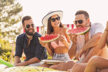 Havuz başında bir yaz partisinde bir grup arkadaş, yüzme havuzunun kenarında oturmuş, soğuk karpuz dilimleri yiyor ve eğleniyorlar.