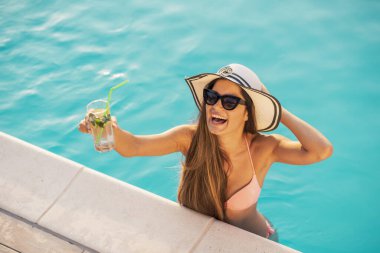 Bikini giyen, güneş gözlüğü takan ve şapka takan çekici genç bir kadın, yüzme havuzunun kenarında duruyor, kokteyller içiyor ve dans ediyor.