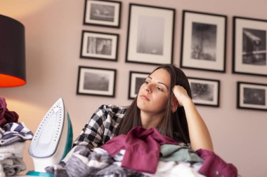 Genç ve yorgun bir kadın ütü masasına yaslanıyor. Etrafı ütüye hazır kırışık kıyafetlerle çevrili.