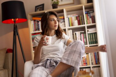 Pijama giyen, oturma odasının penceresinin yanında oturan ve sabah kahvesinin tadını çıkaran genç ve güzel bir kadın.