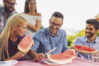 Bir grup arkadaş dışarıda öğle yemeği yiyor, taze karpuz dilimleri yiyor ve eğleniyorlar.