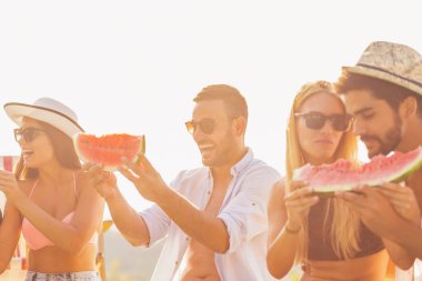 Havuz başında bir yaz partisinde bir grup arkadaş, yüzme havuzunun kenarında oturmuş, soğuk karpuz dilimleri yiyor ve eğleniyorlar.