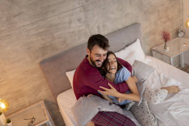 Güzel genç çift pijama giyiyor, yatakta yastık kavgasından sonra gülüyor ve sarılıyorlar.