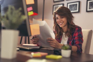 Genç bir kadın, ofisinin masasında oturuyor. İş başarısından sonra elinde neşe dolu belgeler tutuyor.