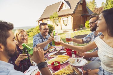 Bir grup genç arkadaş dışarıda barbekü yapıyorlar, masada otururken kadeh kaldırıyorlar.