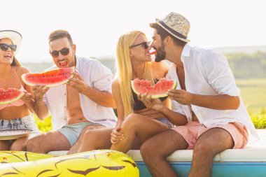 Havuz başında bir yaz partisinde bir grup arkadaş, yüzme havuzunun kenarında oturmuş, soğuk karpuz dilimleri yiyor ve eğleniyorlar.