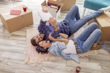 Birbirine aşık çiftin yeni evlerinin zemininde yatıp tablet bilgisayar kullanarak arkadaşlarıyla görüntülü konuşma yapması.