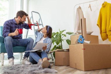 Birbirine aşık bir çift, tablet bilgisayar ve dergiler kullanarak ev dekorasyonu fikirleri ararken kahve içip eğleniyor.