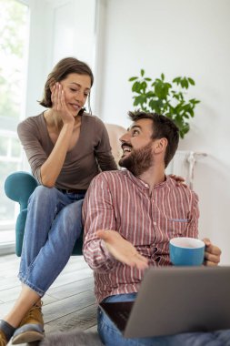 Aşık bir çift yerde oturup kahve içiyor ve dizüstü bilgisayarında ev dekorasyonu fikirleri arıyor.