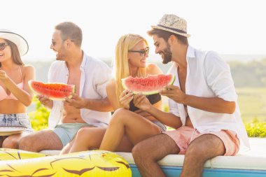 Havuz başında bir yaz partisinde bir grup arkadaş, yüzme havuzunun kenarında oturmuş, soğuk karpuz dilimleri yiyor ve eğleniyorlar.