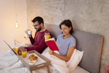 Güzel, genç ve aşık bir çift yatakta kahvaltı ederken internette kitap okuyup sörf yaparken eğleniyor.